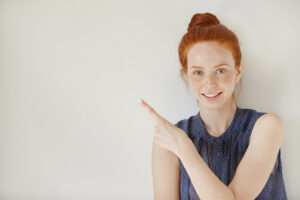Smiling woman with red hair pointing to the side against a plain background.