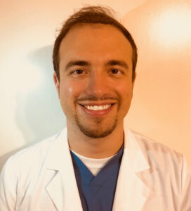 Smiling male doctor in a white coat and blue scrubs.
