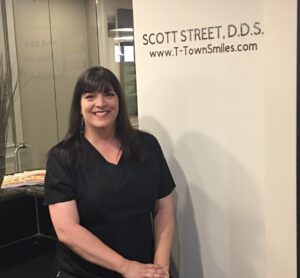 A woman in black scrubs smiling inside a dental office.